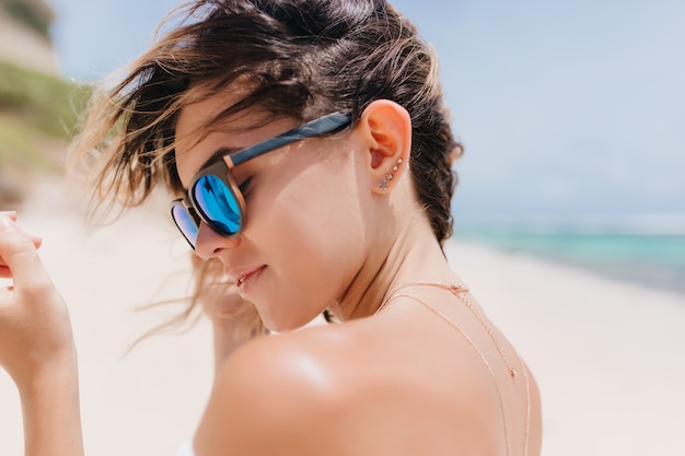 Primo piano sparato di sensuale giovane donna dai capelli scuri in occhiali da sole scintillanti. Bella donna rilassata in piedi sulla spiaggia di sabbia nella calda mattina.