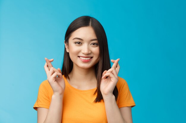 Primo piano speranzosa ragazza asiatica carina con i capelli corti e scuri incrociare le dita buona fortuna, pregando sorridendo ampiamente in attesa di notizie positive, sperando fedelmente di vincere, stare in piedi sfondo blu ottimista eccitato.