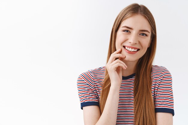 Primo piano tenero, giovane ragazza felice femminile in maglietta a righe, lunghi capelli castani, sorridente a trentadue denti, inclinare la testa divertita, toccare leggermente la guancia con uno sguardo carino, avere una piacevole conversazione