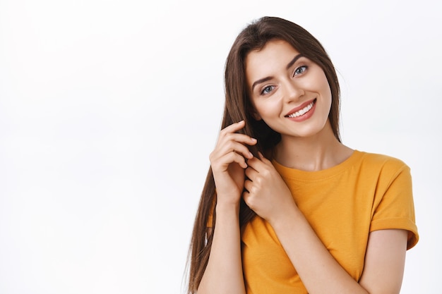 Primo piano tenero, ragazza seducente femminile che si prende cura dei suoi capelli, toccando le ciocche inclinano la testa e sorridendo, soddisfatta del buon prodotto per la cura dei capelli che ha applicato per fare l'acconciatura, sfondo bianco