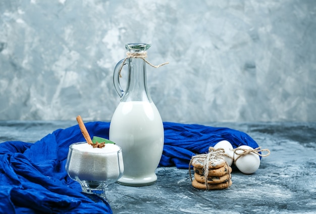Primo piano una brocca di latte con sciarpa blu, gocce di cioccolato e biscotti bianchi e una ciotola di vetro di yogurt su superficie di marmo blu scuro e grigio. orizzontale