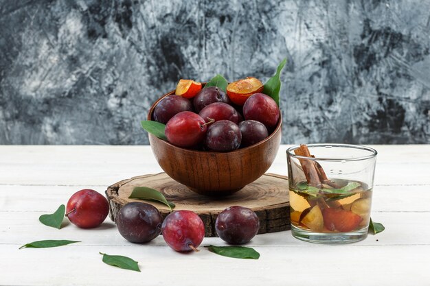 Primo piano una ciotola di prugne sulla tavola di legno con acqua detox e foglie sulla tavola di legno bianca e sfondo di marmo blu scuro. orizzontale