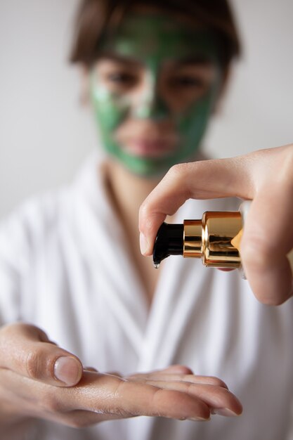 Primo piano, una donna in una veste bianca con una maschera cosmetica verde preme sul distributore di prodotti per la cura della bellezza, sfondo sfocato.