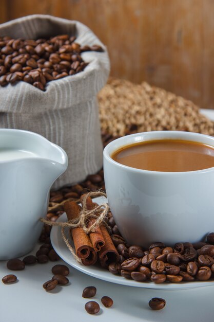 Primo piano una tazza di caffè con chicchi di caffè in un sacco e piattino, latte, cannella secca su sottopentola e superficie bianca. verticale