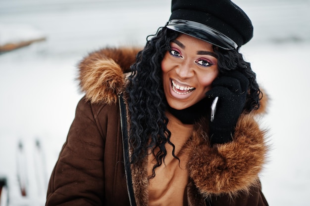 Primo piano volto di donna afroamericana in cappotto di pelle di pecora e cappuccio posato in una giornata invernale su sfondo innevato con il telefono a portata di mano