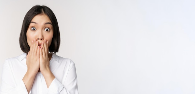 Primo piano volto di donna asiatica ansimante guardando scioccato e senza parole tenendosi per mano vicino alla bocca e fissando la fotocamera impressionato sfondo bianco