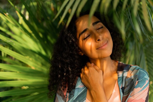 Pritratto di donna adorabile sorridente con i capelli ricci in posa su alberi tropicali e foglie di palma