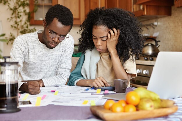 Problema finanziario e concetto di crisi economica. Maschio africano con gli occhiali che ha un'espressione stressata e perplessa, pensando a numerosi debiti, sua moglie infelice seduta accanto a lui e piange