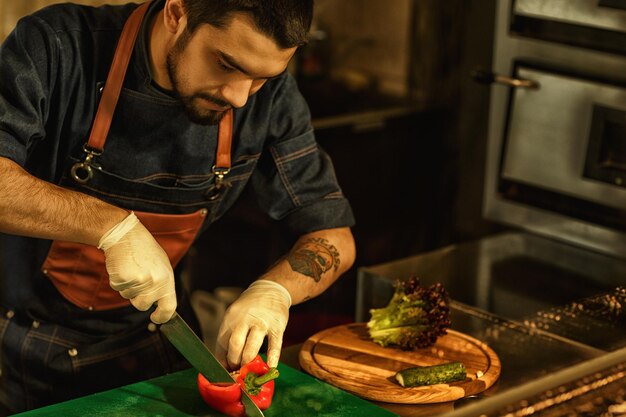 Processo di cottura dello chef di insalata di verdure che taglia la paprika e altre verdure fresche utilizzando un coltello speciale uomo che indossa un grembiule e guanti bianchi sullo sfondo della cucina del ristorante