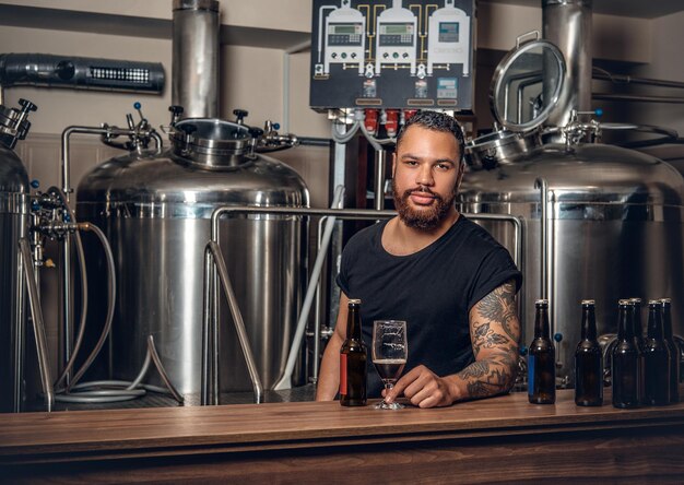 Produttore maschile hipster tatuato con barba nera che presenta birra artigianale nel microbirrificio.