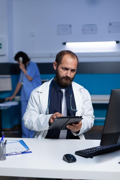 Professionista che utilizza la tecnologia per il sistema sanitario e la pratica medica in ufficio. Dottore che guarda la tavoletta digitale e lo schermo del computer per informazioni di controllo, lavorando fino a tarda notte