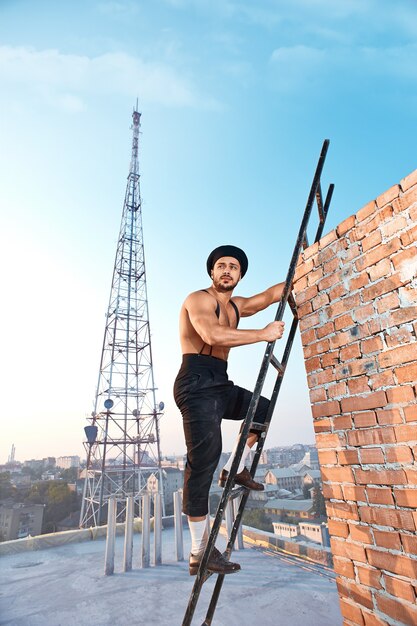 Professionista d'epoca. Muratore muscolare senza camicia che guarda lontano mentre sale una scala in una giornata di lavoro soleggiata