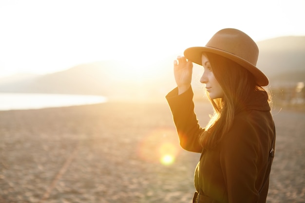 Profilo di affascinante giovane femmina in cappello e cappotto trascorrere una bella serata calda all'aperto