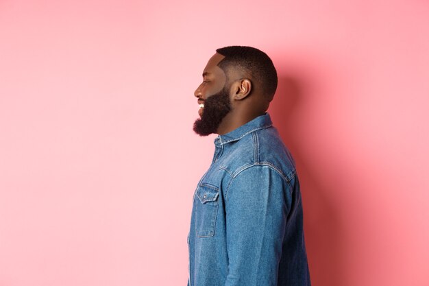 Profilo di bel ragazzo nero barbuto in piedi su sfondo rosa, sorridente e guardando a sinistra lo spazio della copia.