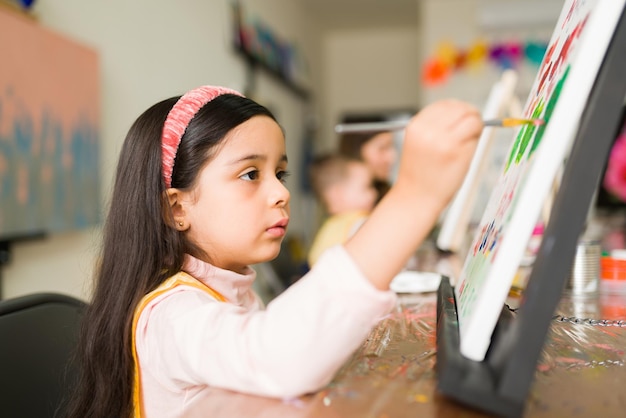 Profilo di una bella ragazza elementare ispanica con un'espressione concentrata che fa un dipinto con un pennello nella sua classe d'arte per bambini