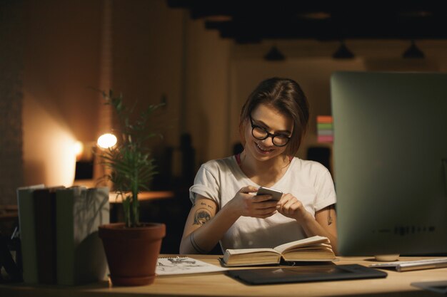 Progettista di signora felice che si siede all'interno di notte chiacchierando per telefono.