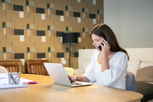 Progettista femminile concentrato che discute del progetto con il cliente sul telefono, seduto al tavolo con il computer portatile e le cianografie e la digitazione