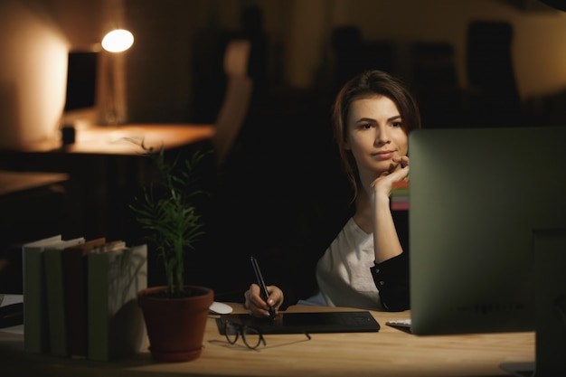 Progettista serio della giovane signora che per mezzo del computer e della tavola dei grafici.