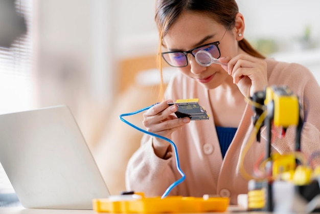 Programmatore femminile asiatico intelligente che impara il cavo della scheda elettronica di codifica del braccio robotico in STEM STEAM cerca di testare il suo braccio robotico autonomo con sensori tramite la piattaforma Arduino a casa