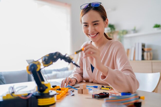 Programmatore femminile asiatico intelligente che impara il cavo della scheda elettronica di codifica del braccio robotico in STEM STEAM cerca di testare il suo braccio robotico autonomo con sensori tramite la piattaforma Arduino a casa