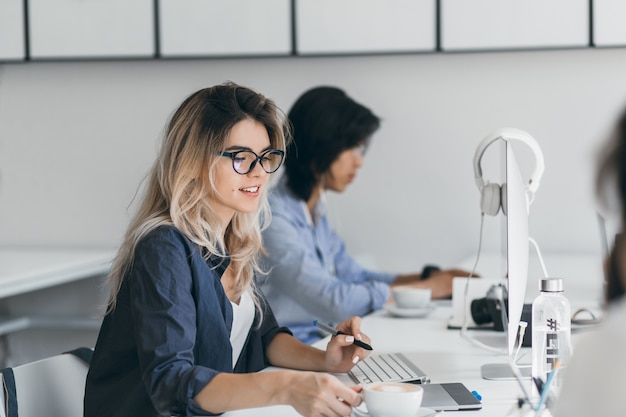 Programmatore femminile dai capelli lunghi che tiene tazza di caffè rilassante dal lavoro. Ritratto dell'interno dello specialista IT europeo seduto al posto di lavoro con il sorriso accanto al giovane asiatico.