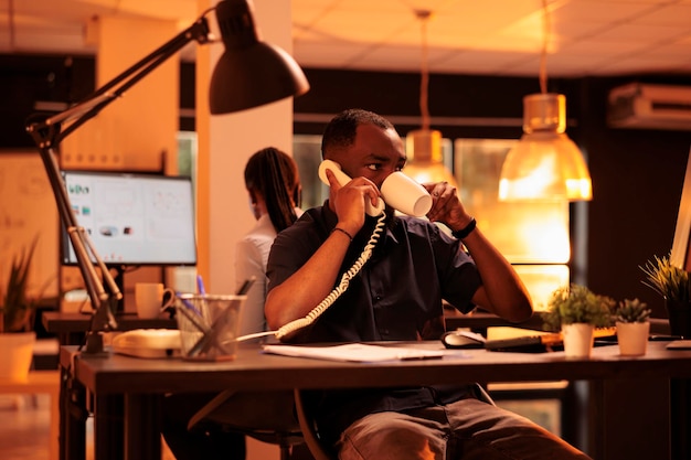 Project manager che utilizza il telefono fisso per avere una conversazione remota durante il tramonto. Chiacchierando sul telefono dell'ufficio con i colleghi per lavorare su rapporti di lavoro e stipulare contratti di scartoffie.