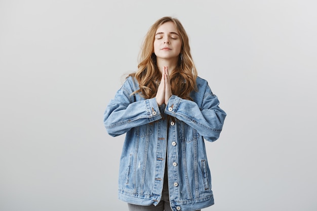 Promettente ragazza bionda in giacca di jeans che esprime desiderio, tenendosi per mano in supplica, pregando