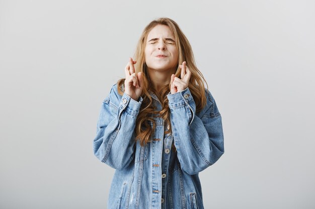 Promettente ragazza bionda in giacca di jeans, esprimere un desiderio, incrociare le dita come pregando