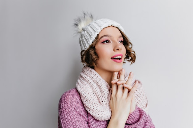 Promettente ragazza riccia dai capelli corti in cappello che guarda lontano. Ritratto del primo piano dell'interno della donna dai capelli scuri piuttosto che posa in sciarpa bianca lavorata a maglia.