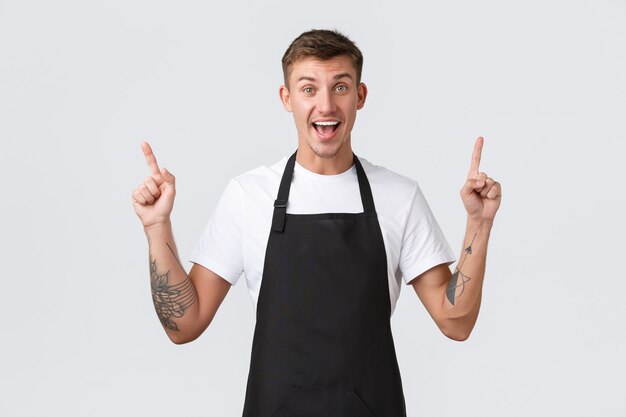 Proprietari di piccole imprese, caffetteria e concetto di personale. Barista entusiasta che invita alla grande apertura, puntando il dito verso il banner e sorridendo eccitato, in piedi sfondo bianco gioioso