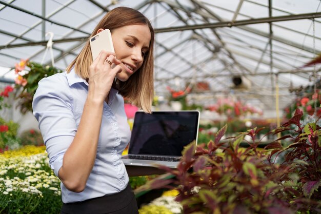 Proprietario di serra sorridente in posa con un computer portatile in mano a parlare al telefono con molti fiori e tetto in vetro.