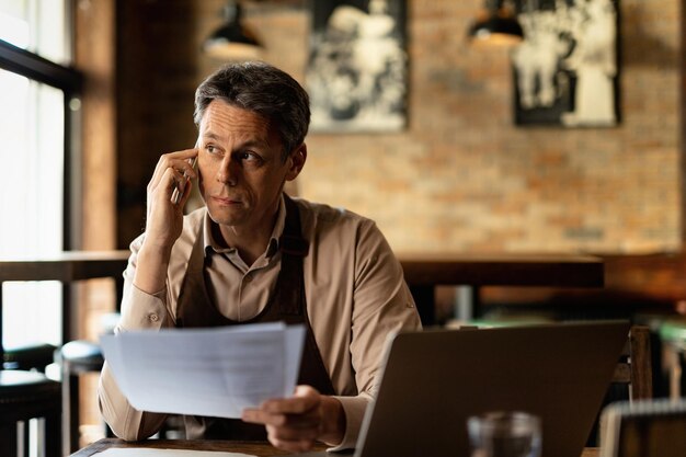 Proprietario di un bar di metà adulto che analizza i documenti e parla al cellulare dopo l'orario di lavoro