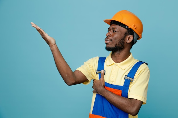 Punti confusi a lato con mano giovane costruttore afroamericano in uniforme isolato su sfondo blu