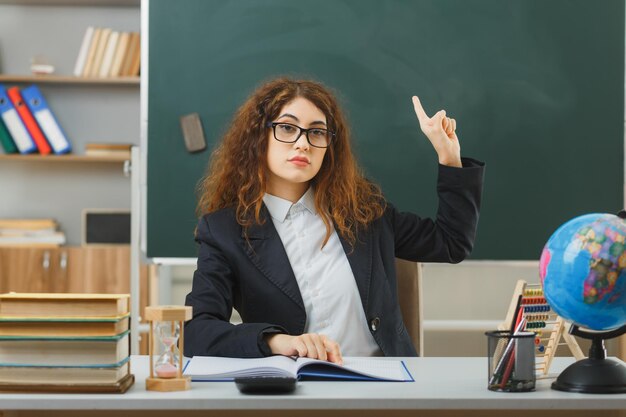 punti fiduciosi su una giovane insegnante femminile che indossa occhiali seduta alla scrivania con strumenti scolastici in classe