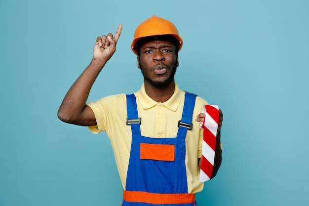 Punti impressionati a tenere il nastro adesivo giovane costruttore afroamericano in uniforme isolato su sfondo blu