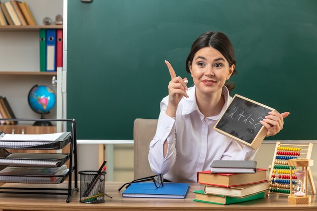 Punti impressionati su una giovane insegnante seduta al tavolo con strumenti scolastici che tengono mini lavagna in classe