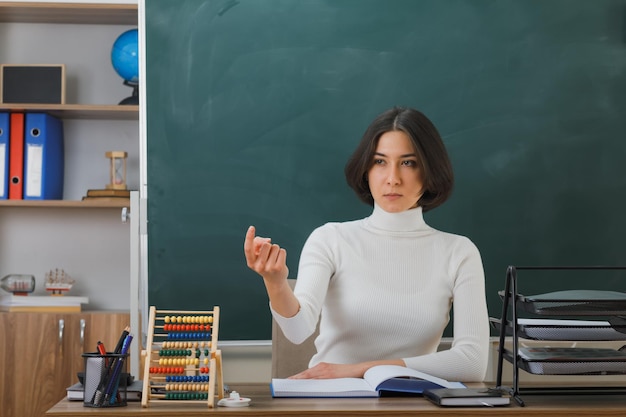 punti severi su una giovane insegnante seduta alla scrivania con gli strumenti scolastici in classe