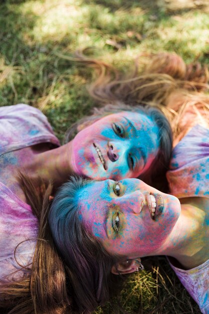 Punto di vista dell&#39;angolo alto delle giovani donne con colore di holi sul loro fronte che si trova sul prato che guarda l&#39;obbiettivo