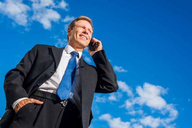 Punto di vista di angolo basso dell&#39;uomo d&#39;affari felice che parla sul telefono cellulare contro il cielo