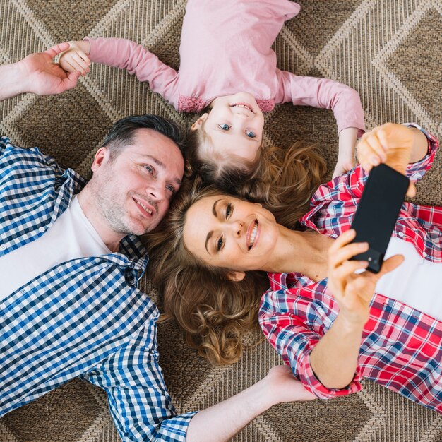 Punto di vista elevato della donna che prende selfie con l&#39;uomo; e figlia nel telefono cellulare mentre giaceva sul tappeto