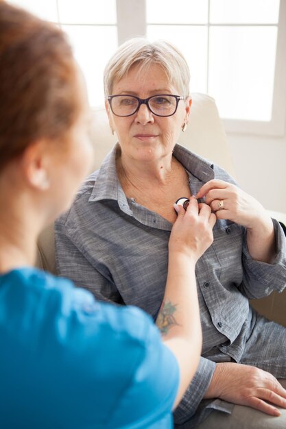 Punto di vista posteriore dell'infermiera femminile che utilizza lo stetoscopio nella casa di cura per ascoltare il battito cardiaco della donna anziana.