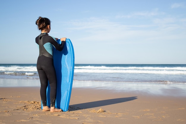 Punto di vista posteriore della bambina con la tavola da surf sulla spiaggia Bambino dai capelli scuri in uniforme da surf in una giornata di sole, guardando l'acqua. Sport, tempo libero, concetto di stile di vita attivo