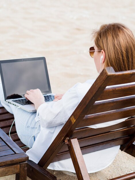 Punto di vista posteriore della donna sulla sedia di spiaggia che lavora al computer portatile