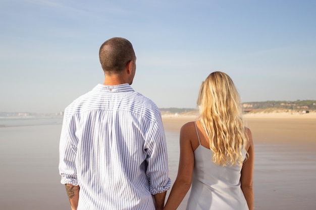 Punto di vista posteriore delle coppie che camminano sulla spiaggia Uomo caucasico con testa rasata e donna in abito casual che si tiene per mano. Amore, vacanza, concetto di affetto