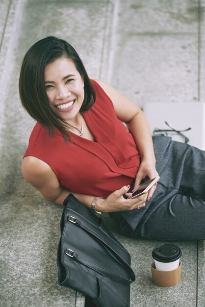 Punto di vista superiore della donna asiatica allegra che dà un sorriso a trentadue denti alla macchina fotografica che riposa sulle scale