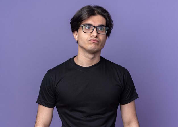 Pursing labbra giovane bel ragazzo che indossa t-shirt nera e occhiali isolati sulla parete viola