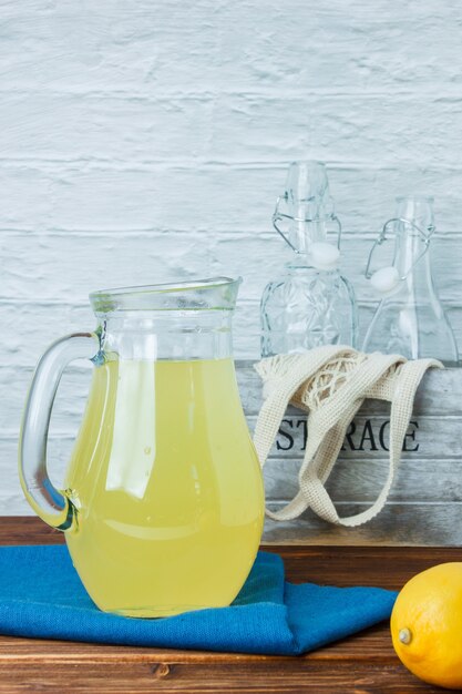 Qualche caraffa di succo di limone con panno blu, bottiglie vuote sulla superficie in legno e bianca, vista laterale.