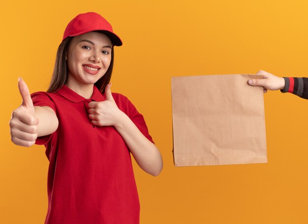 Qualcuno dà il pacchetto di carta alla donna graziosa sorridente di consegna in uniforme che sfoglia con due mani isolate sulla parete arancione con lo spazio della copia