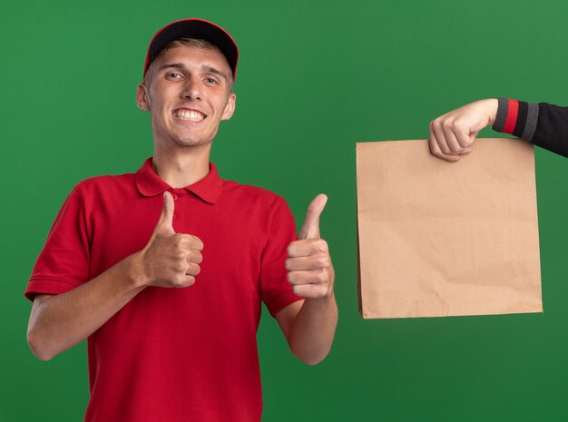 Qualcuno dà un pacco di carta a un giovane ragazzo delle consegne biondo sorridente che fa il pollice in alto con due mani