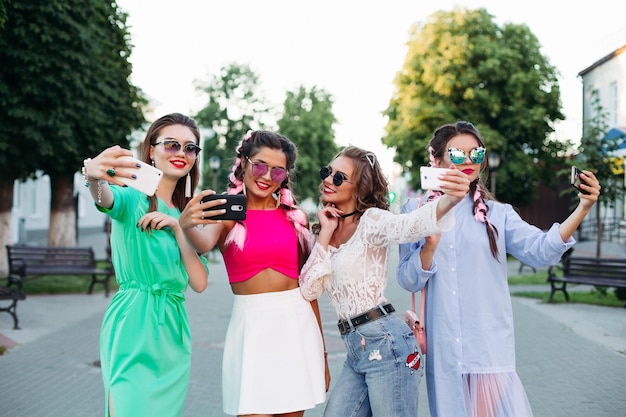 Quattro belle e alla moda migliori amiche con gli occhiali, ognuna in posa facendo selfie per i social network, si divertono. Donne in occhiali da sole dopo lo shopping e il salone di bellezza.